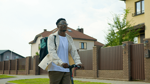 A person with a backpack rides a scooter on a street with houses and fences.