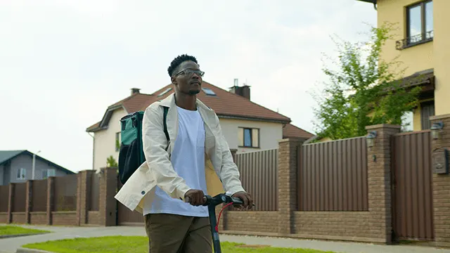 A person with a backpack rides a scooter on a street with houses and fences.