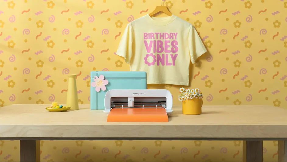 A craft table with a cutting machine, shirt, vase, and box, set against a yellow patterned wall.