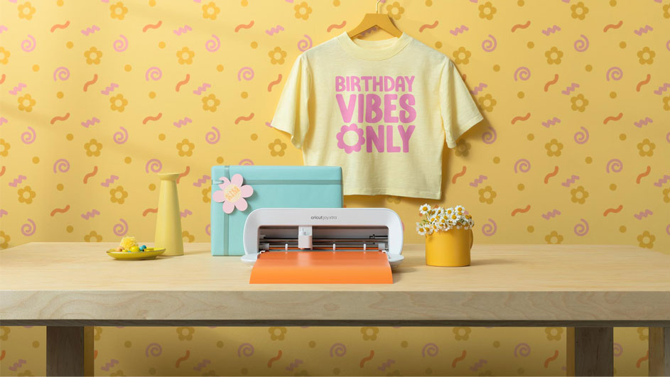 Een geel T-shirt met de tekst "Birthday Vibes Only" hangt boven een tafel met een snijmachine, bloemen en decoraties.