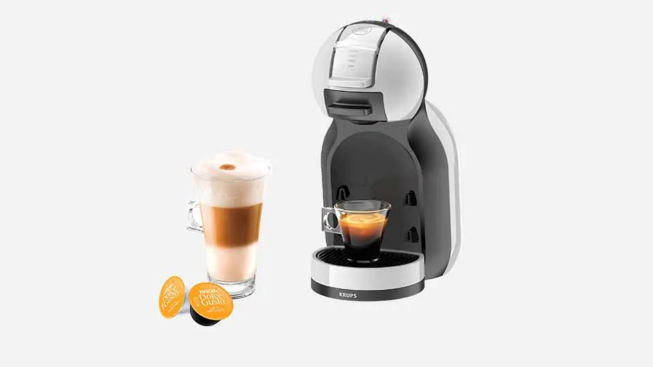 Een koffiezetapparaat staat naast een glas gelaagde koffie en twee koffiecapsules op een witte achtergrond.