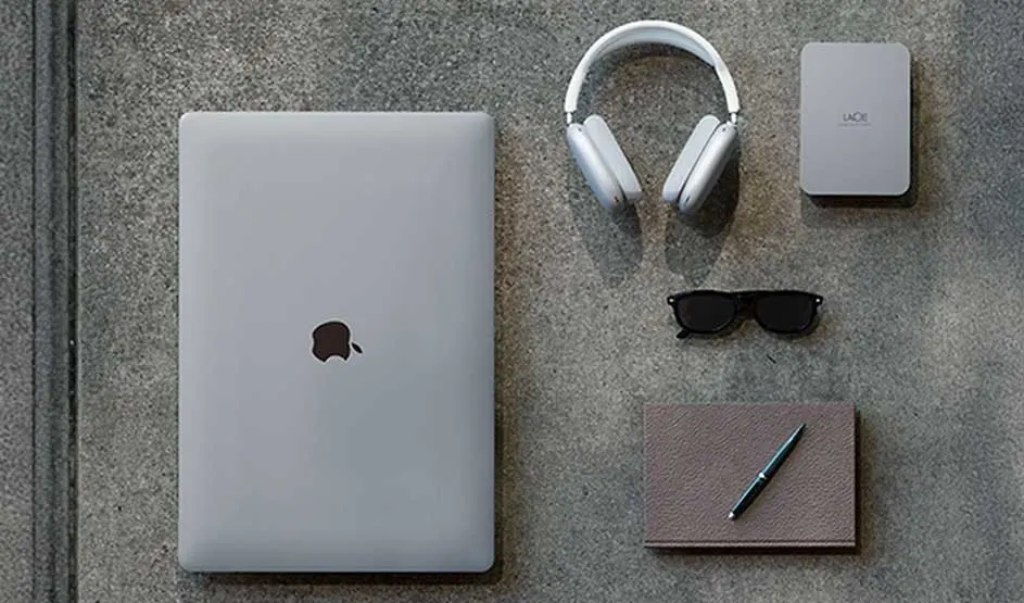 An Apple MacBook, LaCie hard drive, headphones, sunglasses, a notebook, and a pen are neatly arranged on a grey surface.