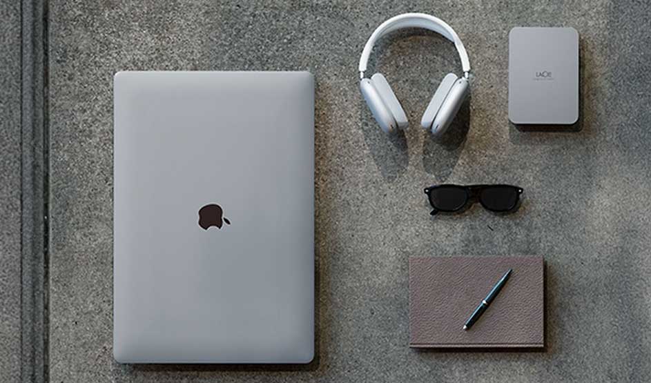 Op de afbeelding zie je een Apple laptop, LaCie harde schijf, koptelefoon, zonnebril, notitieboek en pen netjes op een grijze ondergrond.