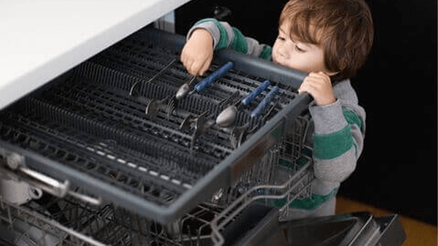 Een kind staat bij een open vaatwasser met een rek vol zilverwerk, waaronder lepels en vorken met blauwe handvatten.