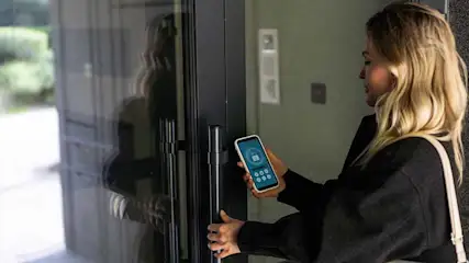 Een vrouw opent een zwarte deur met een zilveren handgreep, met een telefoon met een app om de deur te ontgrendelen.
