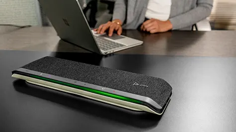 Poly speakerphone met groene statuslijn staat op donker bureau, terwijl iemand achter een open laptop aan een videogesprek werkt.