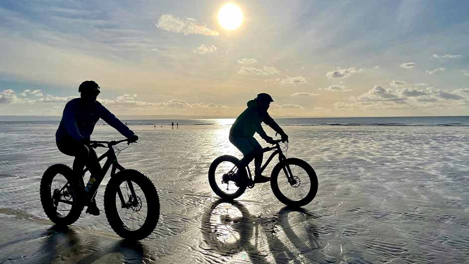 Twee mensen fietsen op een strand met een blauwe lucht en de zon op de achtergrond.