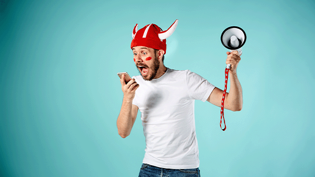 Man met rode vikinghelm en vlaggen op wangen, kijkt opgewonden naar telefoon en houdt megafoon vast tegen blauwe achtergrond.