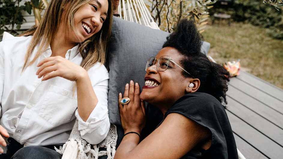 Twee vrouwen lachen in een hangmat met een grijs kussen. Een vrouw draagt een wit overhemd, de ander een zwart shirt en een ring.