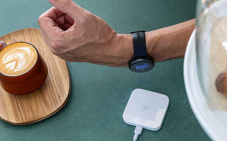 A person pays contactless with their Garmin smartwatch at a payment terminal, next to a cappuccino in a café.