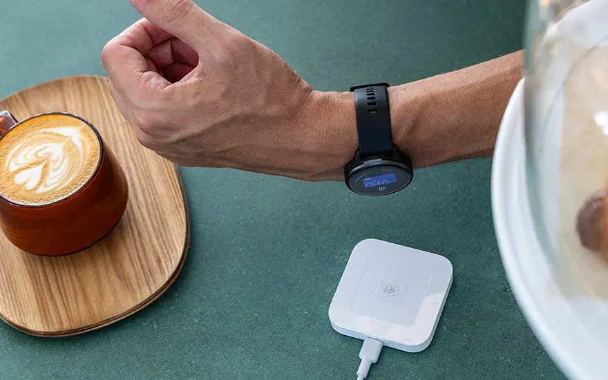 Een persoon betaalt contactloos met zijn Garmin smartwatch bij een betaalterminal, naast een cappuccino in een café.