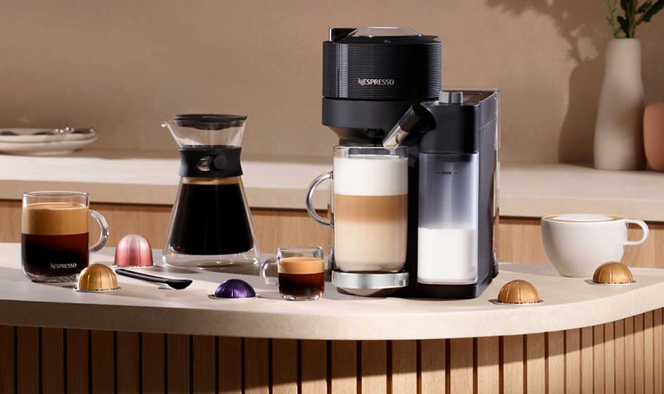 Een Nespresso-koffiemachine met melkkan staat op een aanrecht, naast glazen koffie, capsules, een karaf en een witte mok.