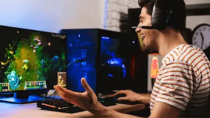 A person with a headset plays a video game on a desktop computer with a monitor and a blue light-up computer tower on a desk.