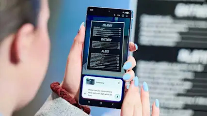 Een vrouw houdt een Samsung-smartphone verticaal en scant een restaurantmenu, terwijl AI-tekst onderaan suggesties of vertaling toont.