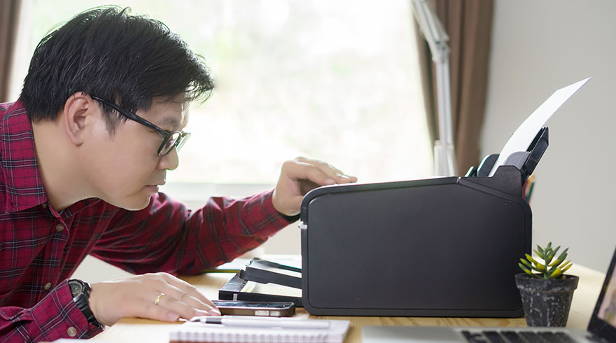 Een persoon met een geruit rood hemd staat bij een printer op een bureau met een laptop, notitieboek en plant.