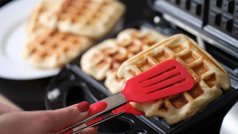 Close-up van een wafelijzer waarin verse wafels worden gebakken; een hand gebruikt een rood siliconen spateltje om een wafel uit het apparaat te halen, met reeds gebakken wafels op een wit bord op de achtergrond.