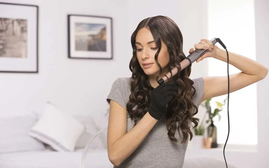 Vrouw in grijze top stylet haar donkere krullen met een krultang, draagt een beschermende handschoen in een lichte kamer.