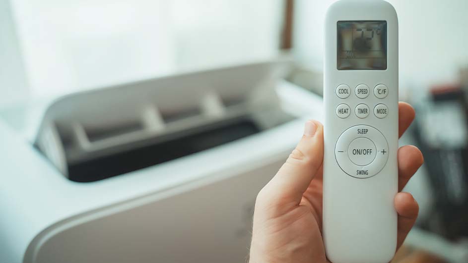 Een hand houdt een witte afstandsbediening vast met een scherm en knoppen, voor een witte airconditioner.