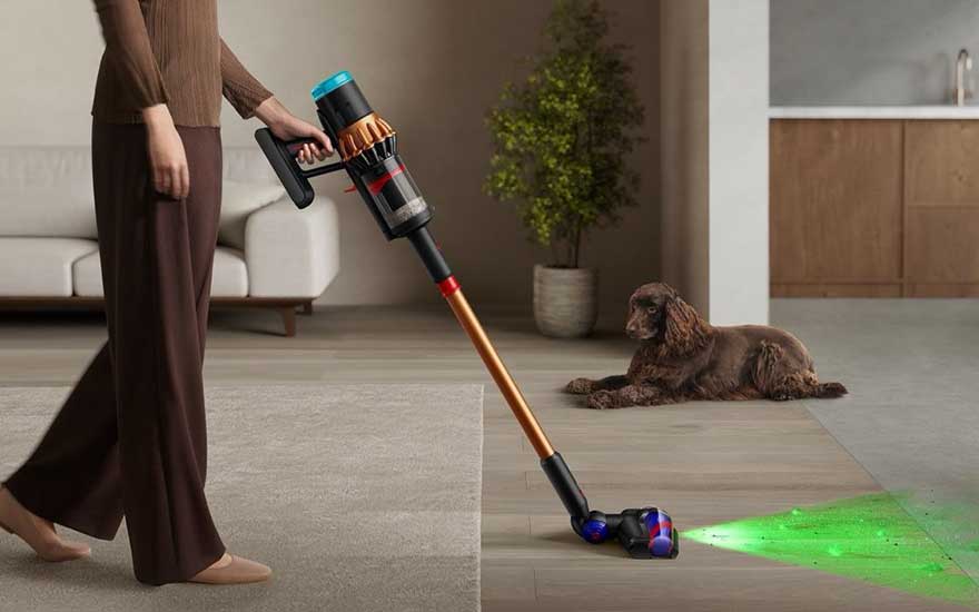 A woman is vacuuming a rug with a stick vacuum featuring a green laser, while a dog is lying on the wooden floor nearby.