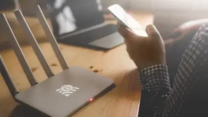 Een grijze router met vier antennes staat op een houten tafel, naast een laptop en iemand met een telefoon.
