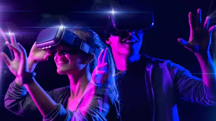Two people wearing VR headsets are smiling and reaching out their hands, fully immersed in a vibrant, virtual world.