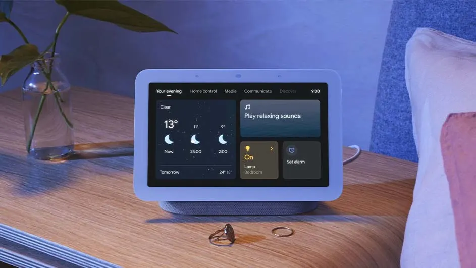 Een slim scherm toont het weer, de tijd en bedieningselementen voor een lamp en alarm, op een houten tafel naast een vaas en sieraden.