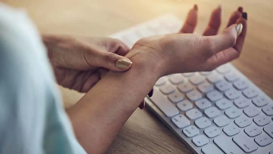 Een hand masseert de pols boven een wit toetsenbord op een houten bureau.