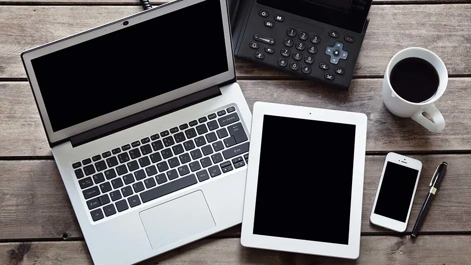 Een laptop, tablet, telefoon, rekenmachine, pen en een kop koffie liggen op een houten tafel.
