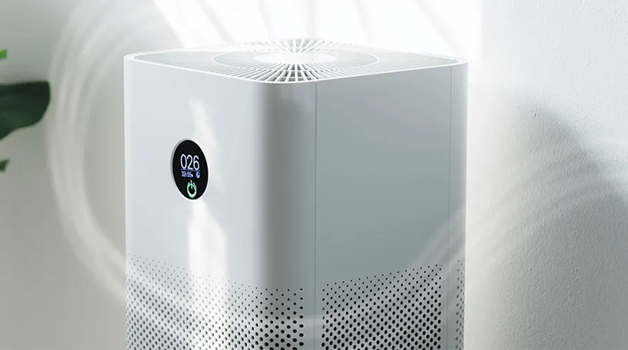 A white, rectangular air purifier with a digital display showing "026" sits against a light wall. The bottom has small holes.