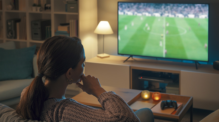 Vrouw kijkt naar een voetbalwedstrijd op een grote televisie in een woonkamer met een lamp en een salontafel.