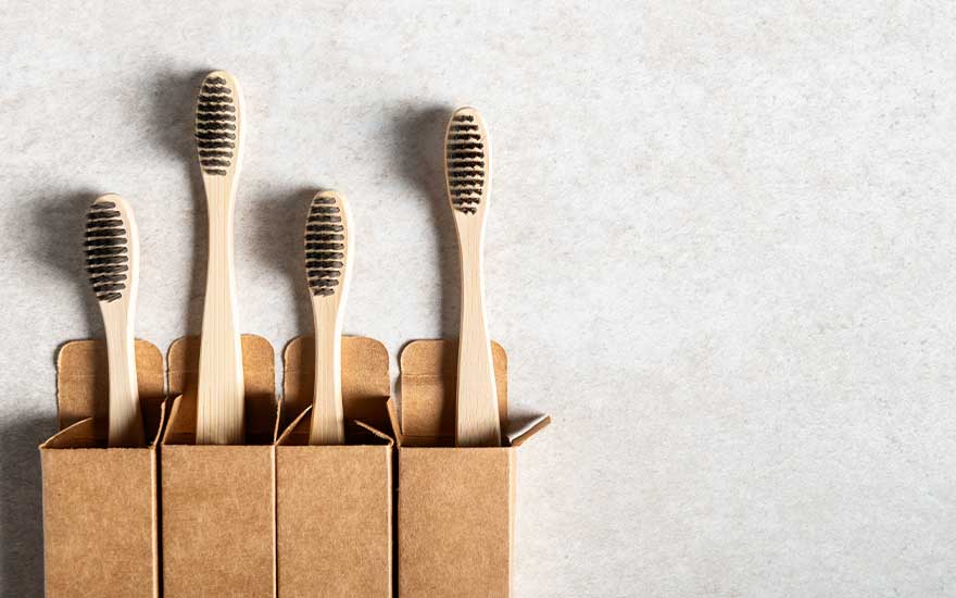 Four bamboo toothbrushes with dark bristles stand in simple cardboard boxes, set against a light, neutral textured background.