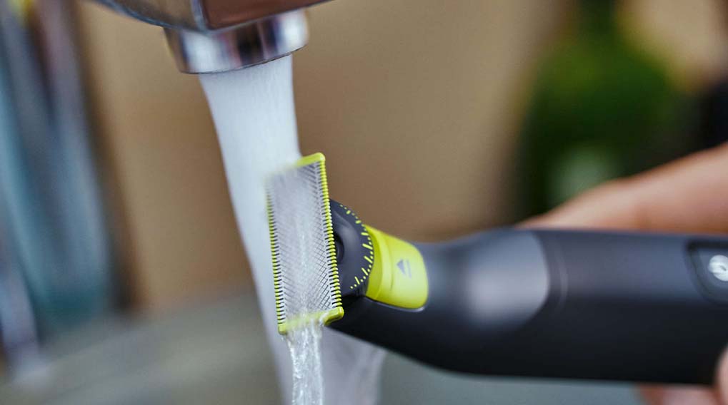 Een zwart-groene trimmer wordt schoongespoeld onder een zilveren kraan met stromend water, de achtergrond is vaag.