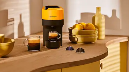 Espressomachine naast kopjes, capsules en citroenen op een houten aanrecht. Een fles staat achter de citroenen.