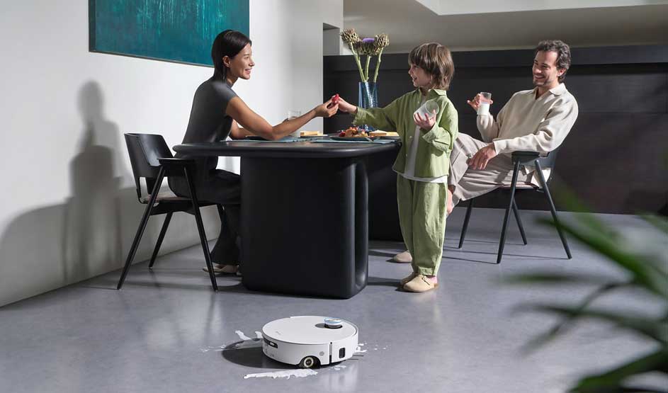 Een gezin zit aan tafel; een kind geeft iets aan een vrouw, terwijl een witte robotstofzuiger op de vloer door een gemorste plas rijdt.