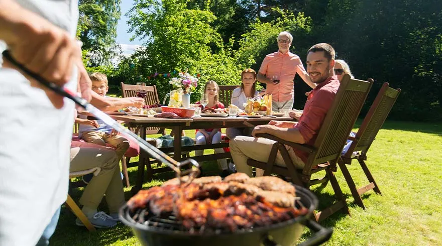 Een buitenscène met een groep mensen rond een houten tafel vol eten, met een zwarte barbecue op de voorgrond.