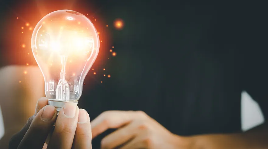 Een hand houdt een gloeilamp vast die fel oranje licht uitstraalt tegen een donkere achtergrond.