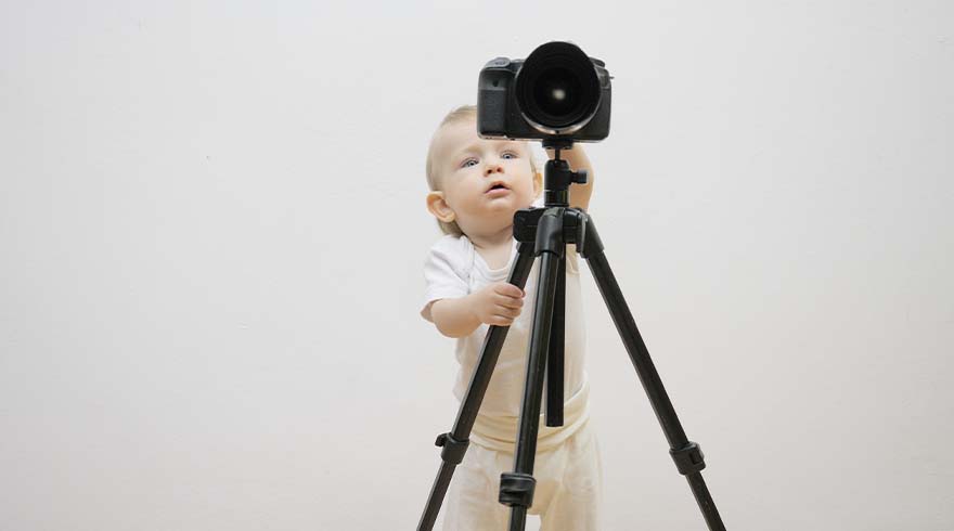 Een baby in witte kleding staat voor een zwart statief met een zwarte camera erop, tegen een witte achtergrond.