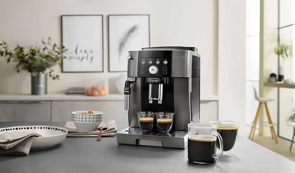 Op de afbeelding staat een zwarte De’Longhi espressomachine op een aanrecht, met vier glazen koffie en wat servies ernaast.