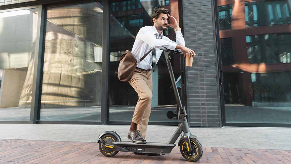 A person on a scooter, wearing a shirt, tie, and slacks, with a shoulder bag and a cup, outdoors on a sidewalk.