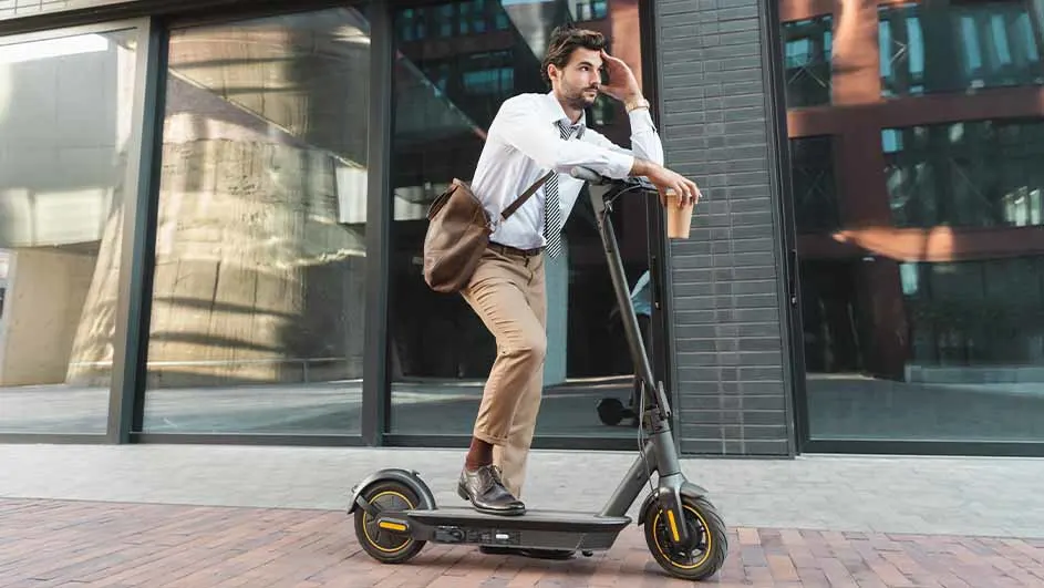 Een man in overhemd en broek staat op een elektrische scooter met een tas en een beker in de hand.