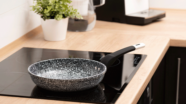 Een zwarte kookplaat met een zwarte pan met een wit gespikkelde binnenkant. Een plant en een koffiezetapparaat staan op het aanrecht.