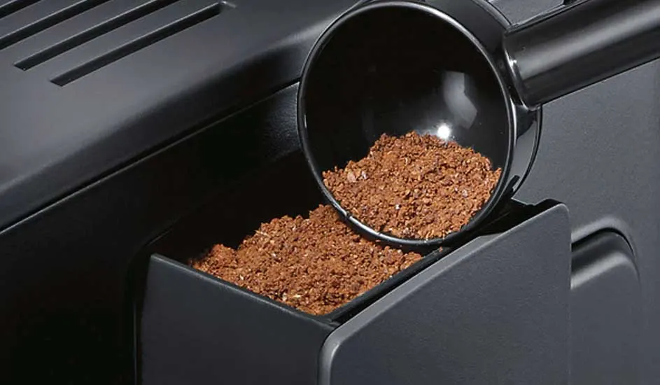 Een zwarte lepel schept versgemalen koffie uit een bakje van een koffiemachine, klaar om gebruikt te worden voor een kop koffie.