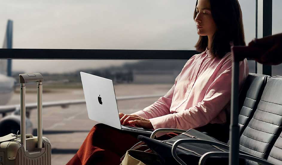 Persoon in roze blouse werkt op MacBook in luchthavenwachtruimte met koffer ernaast en uitzicht op vliegveld.