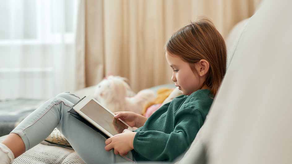 Meisje zit op een bank met een tablet, gekleed in een groene trui en een lichtblauwe broek. Op de achtergrond een gordijn.