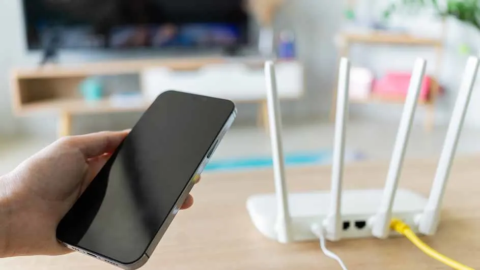 Een hand houdt een zwarte smartphone vast naast een witte router met drie antennes op een houten tafel, met een tv op de achtergrond.