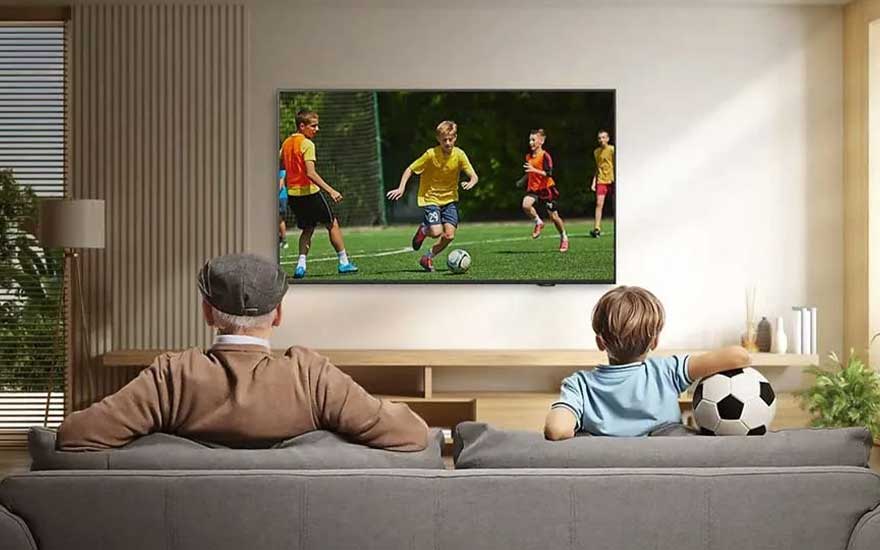 An elderly man and a boy with a football sit on a sofa watching a children’s football match on a large television.