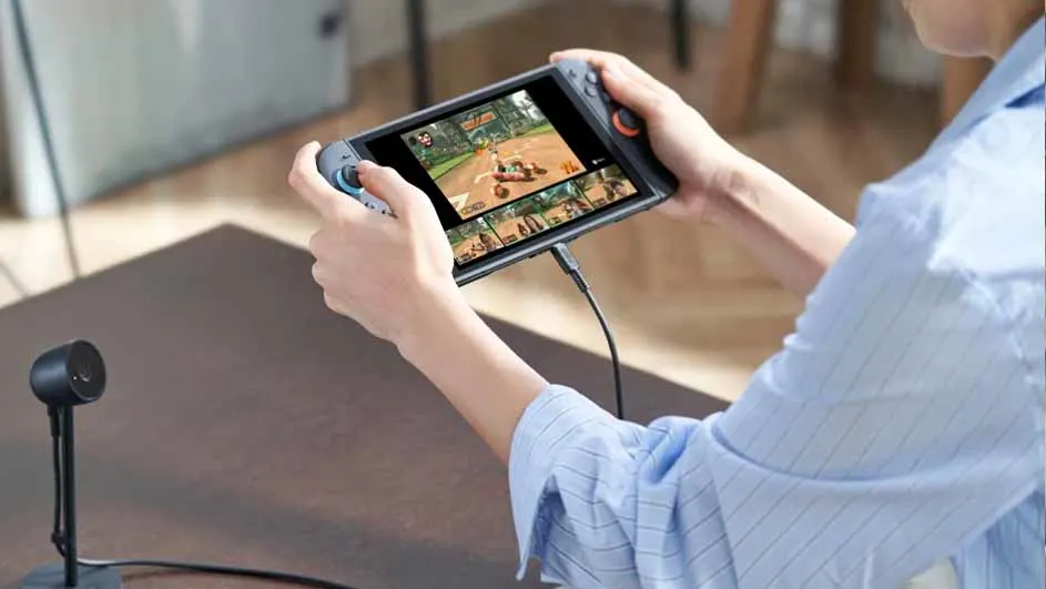 Een persoon speelt een racespel op een handheld apparaat met een zwarte kabel. Een zwarte microfoon staat op een bruine tafel.