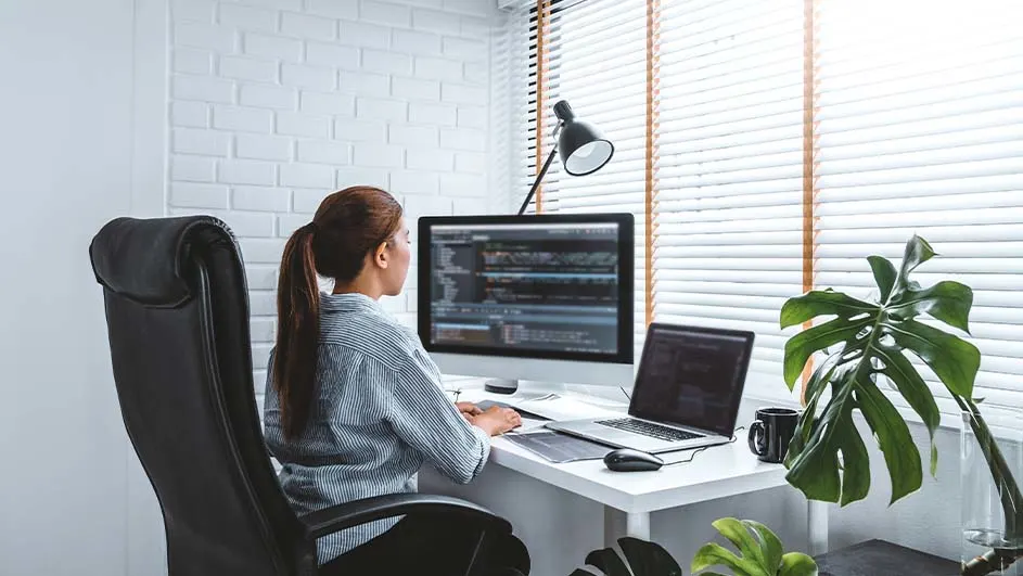 Vrouw zit aan bureau met computer en laptop in lichte kamer met planten en jaloezieën.