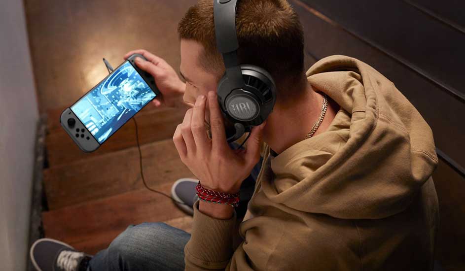 A young man on wooden stairs wears large JBL headphones, leaning forward as he plays an intense game on a handheld console.