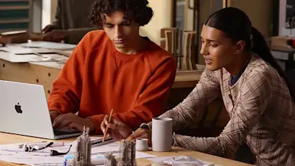 Two students collaborate at a wooden workbench, one sketching designs while the other types on a laptop beside small architectural models.
